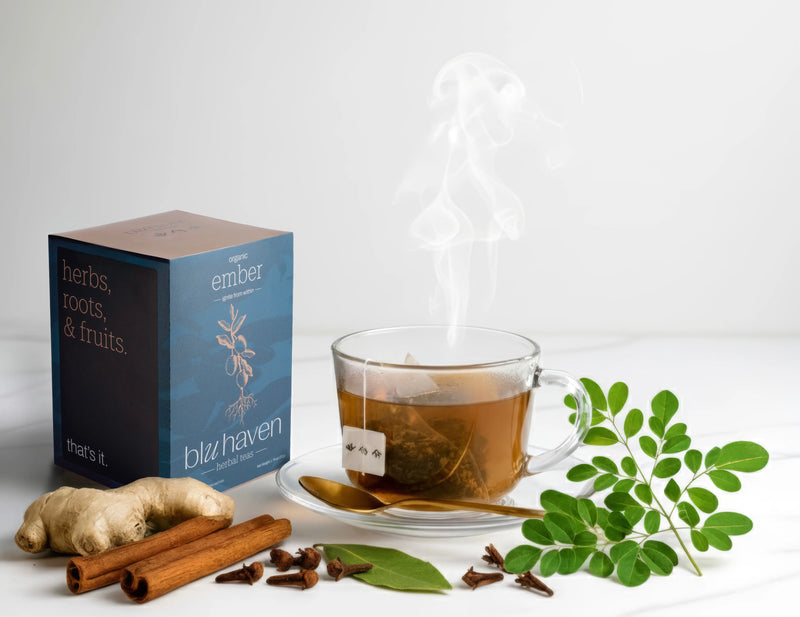 A box of Blu Haven 'Ember' organic herbal tea next to a steaming glass cup of tea with a pyramid sachet. Ingredients like fresh ginger, cinnamon sticks, cloves, and moringa leaves are scattered on a white marble surface.