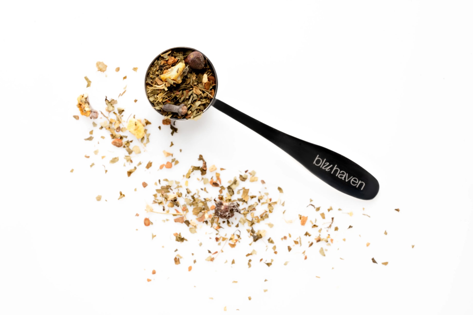 Black spoon filled with textured loose leaf herbal tea blend spilling onto a white surface.